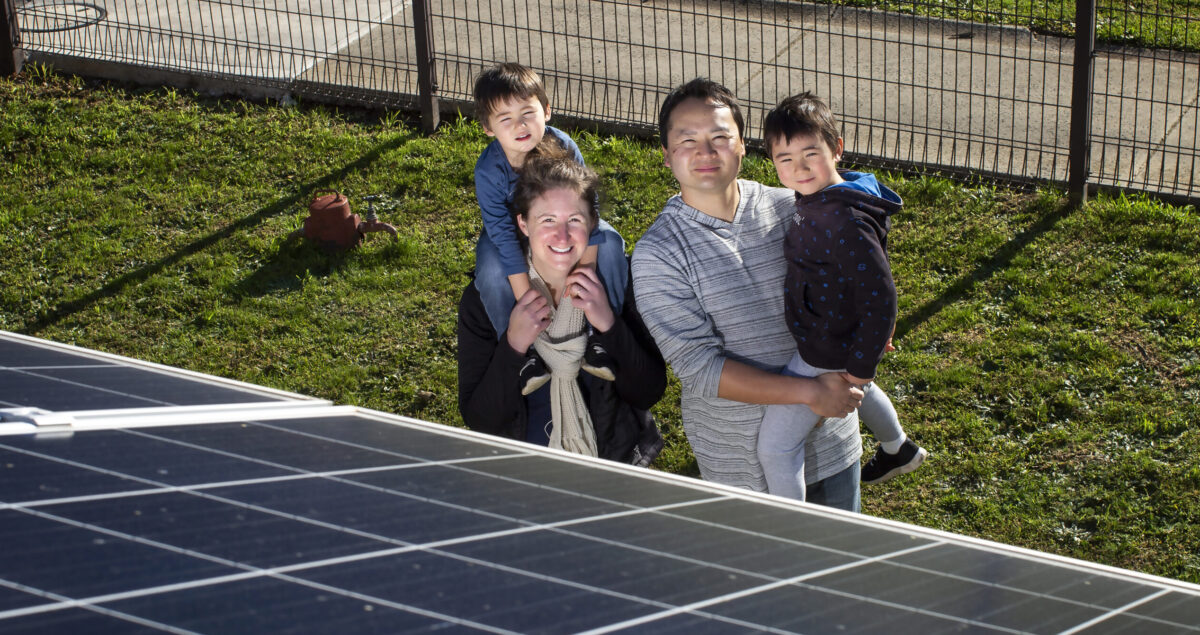 Smiling couple with two young children standing outdoors near solar panels, with one child on a parent’s shoulders and the other being carried.