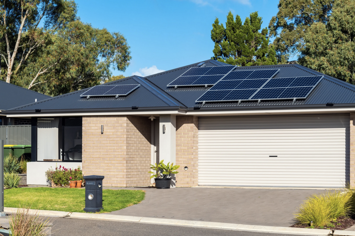 Single-storey brick house with solar panels installed across the roof, viewed from the street with trees in the background.