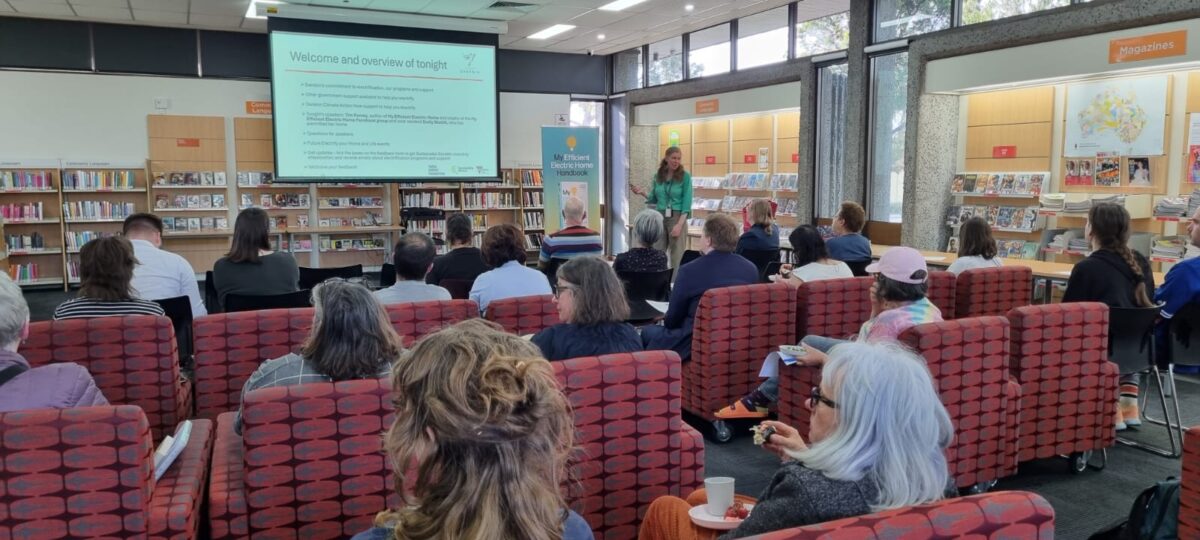 Audience seated in a library listening to a presenter standing beside a screen with slides during a community electrification information session.